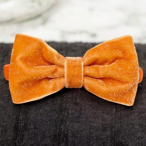 Puede incluir: Una pequeña pajarita de terciopelo naranja con un brillo sutil. La pajarita está atada con un solo lazo y tiene un diseño simple y elegante.