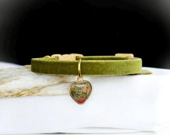 May include: Olive green velvet pet collar with a gold-colored buckle and a heart-shaped charm. The charm features a stone with red and green inclusions. The collar is displayed on a white surface.