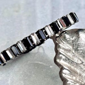 May include: A black and clear crystal bracelet with rectangular shaped stones. The bracelet is on a silver leaf background.