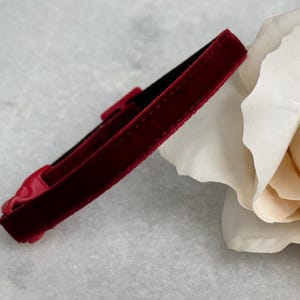 May include: A close-up of a red velvet cat collar with a buckle closure. The collar is lying on a white surface next to a white rose.