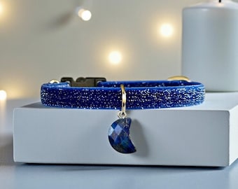 May include: A blue velvet pet collar featuring a glittery texture and a black plastic buckle. A crescent moon-shaped charm with a faceted, dark blue stone finish hangs from a gold-toned metal ring attached to the center of the collar band.