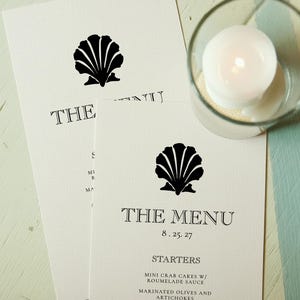 May include: Off-white menu cards with a black seashell design and the text "THE MENU" with the date 8.25.27. A lit white candle sits in a clear glass holder. The menu lists starters including mini crab cakes and marinated olives.
