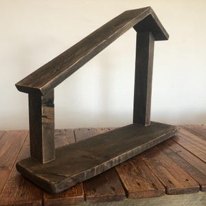 May include: A rustic, dark brown wooden shelf with a house-shaped design. The shelf features a flat base, two vertical supports, and a slanted roof section, all constructed from weathered wood. The design is simple and minimalist.