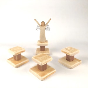 May include: A wooden angel figurine with wire wings stands atop a small, light-colored wooden pedestal. Three additional pedestals with square tops and brown wooden bases are arranged nearby. The pedestals are made of light-colored wood.
