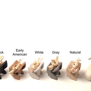 May include: A collection of small, decorative figurines in various colors, including black, Early American, white, gray, and natural. Each figurine features a small bundle of straw. A larger figurine depicts a woman holding a child.