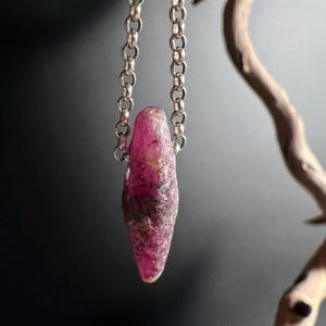 Puede incluir: Colgante de piedra de rubí en bruto, suspendido de una cadena de plata. El rubí, de color rosa intenso, presenta texturas naturales. La cadena está compuesta por pequeños círculos enlazados. El colgante mide unos 3 cm de largo.
