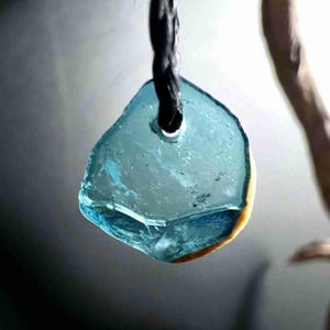 Pu&ograve; includere: Un ciondolo azzurro di forma irregolare con un cordino nero. Il ciondolo ha un foro nella parte superiore per il cordino e un bordo marrone dorato. Il ciondolo &egrave; sospeso su uno sfondo sfocato.