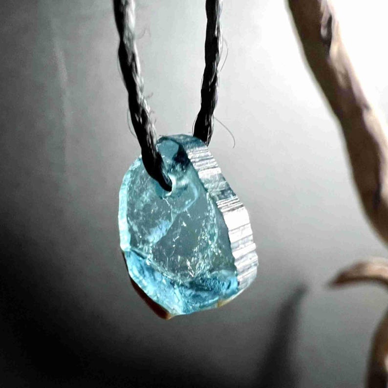 Pu&ograve; includere: Un pendente azzurro, di forma irregolare, realizzato in pietra traslucida, sospeso a un cordino nero. La pietra ha un bordo sfaccettato e un aspetto naturale e grezzo. Lo sfondo &egrave; grigio tenue.