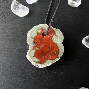 May include: A red and green agate pendant with a black cord. The pendant is irregularly shaped and has a natural, rough edge.