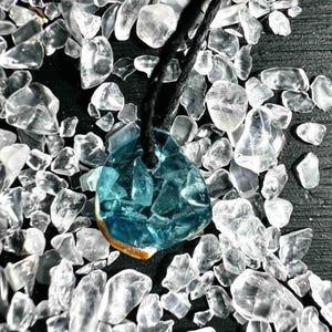 Pu&ograve; includere: Una collana con pendente artigianale con un pendente in vetro azzurro a forma di goccia. Il pendente &egrave; composto da pezzi di vetro fusi ed &egrave; sospeso a un cordino nero. Lo sfondo &egrave; pieno di pezzi di vetro chiari e irregolari.