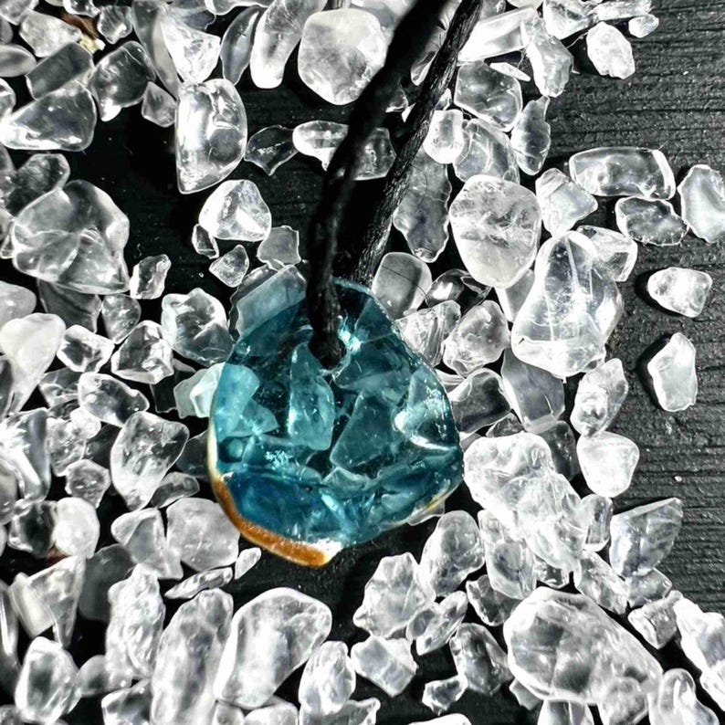 Pu&ograve; includere: Una collana con pendente artigianale con un pendente in vetro azzurro a forma di goccia. Il pendente &egrave; composto da pezzi di vetro fusi ed &egrave; sospeso a un cordino nero. Lo sfondo &egrave; pieno di pezzi di vetro chiari e irregolari.