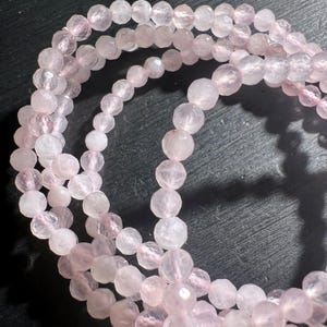 Peut inclure: Un brin de perles de quartz rose, facettées et polies, avec une teinte rose clair. Les perles sont disposées en cercle, créant un effet de superposition.