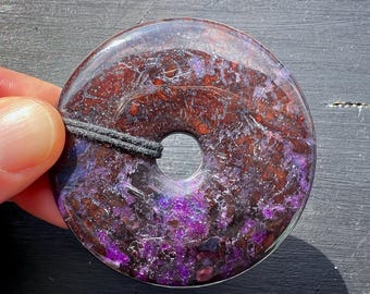 Puede incluir: Un colgante redondo de piedra preciosa de color púrpura y marrón con un agujero en el centro para un cordón. El colgante está hecho de una piedra pulida con un acabado brillante e iridiscente.