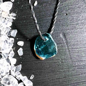 Pu&ograve; includere: Una collana con pendente turchese con un cordino scuro. Il pendente ha una forma irregolare con un bordo marrone. La collana &egrave; esposta su una superficie di legno scuro, con cristalli bianchi sparsi intorno.