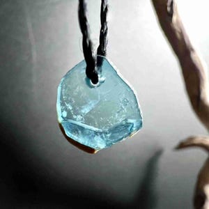 Pu&ograve; includere: Un ciondolo azzurro chiaro, di forma irregolare, forse in vetro o cristallo, &egrave; appeso a un cordino nero. Il ciondolo ha un aspetto naturale e organico con una consistenza leggermente ruvida e un piccolo foro nella parte superiore per il cordino.