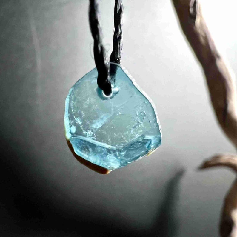Pu&ograve; includere: Un ciondolo azzurro chiaro, di forma irregolare, forse in vetro o cristallo, &egrave; appeso a un cordino nero. Il ciondolo ha un aspetto naturale e organico con una consistenza leggermente ruvida e un piccolo foro nella parte superiore per il cordino.