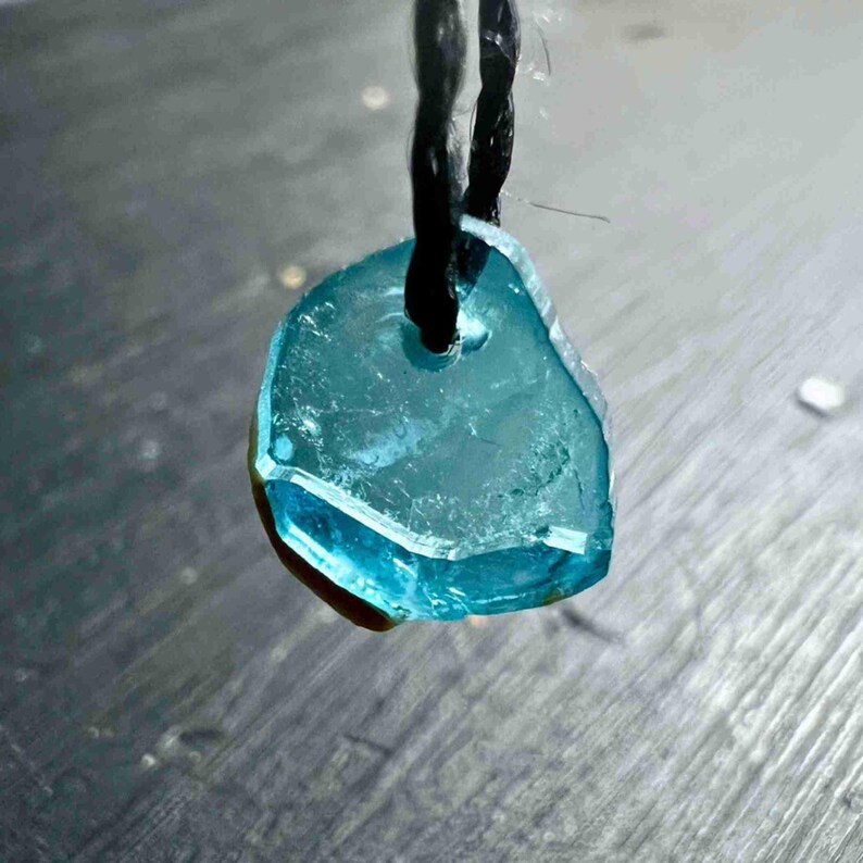 Pu&ograve; includere: Un ciondolo azzurro, di forma irregolare, realizzato in un materiale traslucido, forse una pietra preziosa o vetro marino. &Egrave; sospeso a un cordino nero. Il ciondolo ha un foro nella parte superiore per il cordino.