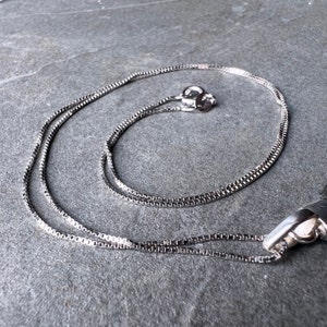 Può includere: Una collana a catena d'argento con chiusura a scatola. La catena è composta da piccoli anelli quadrati.