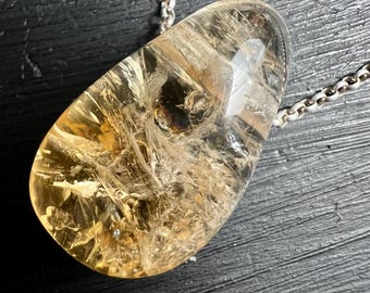 Puede incluir: Un colgante en forma de lágrima hecho de cuarzo citrino amarillo dorado translúcido con fracturas internas visibles e inclusiones en forma de aguja, suspendido de una cadena de metal plateado con pequeños eslabones circulares.