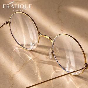 May include: Gold-rimmed eyeglasses with clear lenses. The round frames have a delicate design, with thin gold arms and nose pads. The brand name "ERATIQUE EYEWEAR" is printed at the top. The glasses are set against a neutral background.