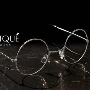 May include: Silver-rimmed eyeglasses with round lenses, casting a shadow on a dark marble surface. The brand name "ERATIQUE EYEWEAR" is displayed in white. The glasses have a minimalist design.