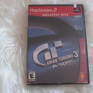 May include: A PlayStation 2 video game case for Gran Turismo 3: A-spec. The case has a dark blue background with the game's logo and the PlayStation 2 Greatest Hits logo. The game is rated "E" for Everyone.