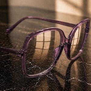 May include: Translucent purple square-framed eyeglasses with clear lenses. The frame features the text ERATIQUE EYEWEAR in the upper left corner. The glasses have a glossy finish and thin temples, showcasing a modern design with a bold, oversized shape.
