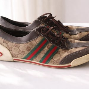 Puede incluir: Par de zapatillas marrones y beige con el logo de Gucci. Las zapatillas presentan el ic&oacute;nico patr&oacute;n de Gucci, cordones marrones y rayas rojas y verdes. La suela es blanquecina.