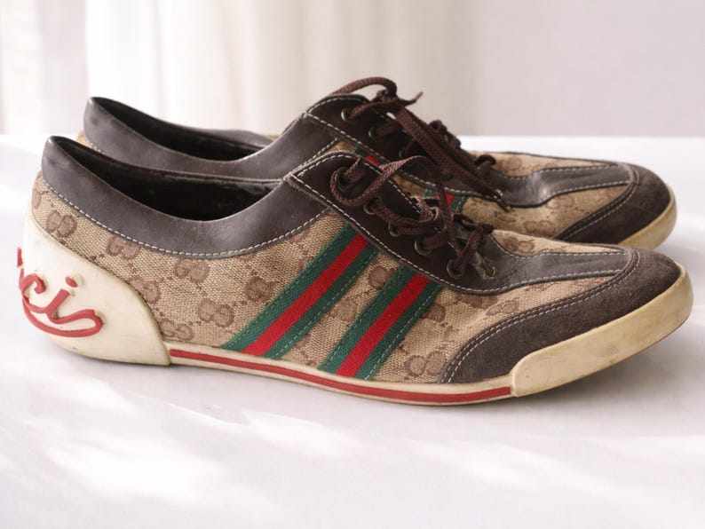 Puede incluir: Par de zapatillas marrones y beige con el logo de Gucci. Las zapatillas presentan el ic&oacute;nico patr&oacute;n de Gucci, cordones marrones y rayas rojas y verdes. La suela es blanquecina.