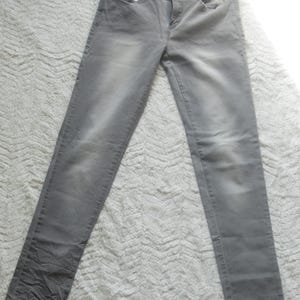 May include: A pair of gray denim jeans. The jeans have a classic five-pocket design, a button and zipper closure, and a faded appearance. The fabric appears to be a medium-weight denim. The jeans are laid flat on a textured surface.