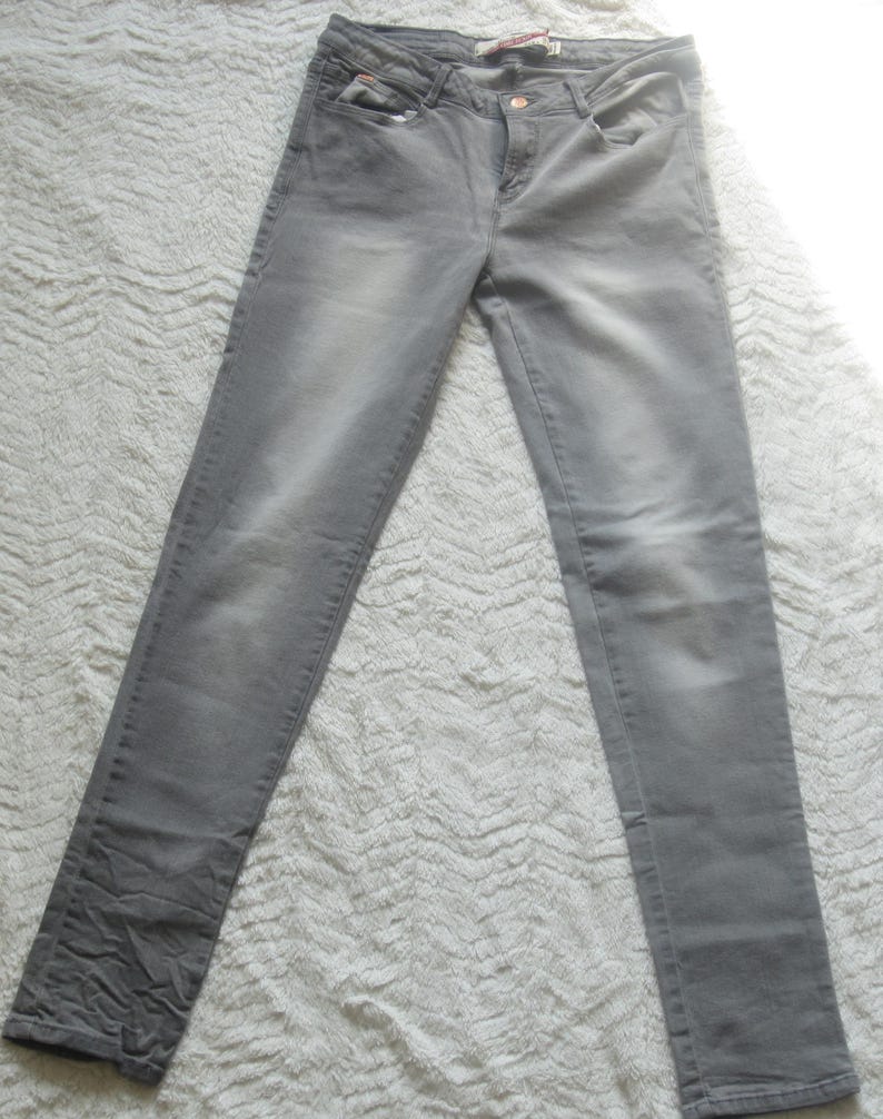 May include: A pair of gray denim jeans. The jeans have a classic five-pocket design, a button and zipper closure, and a faded appearance. The fabric appears to be a medium-weight denim. The jeans are laid flat on a textured surface.