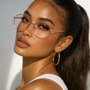 May include: A woman wearing round, gold-rimmed eyeglasses and gold hoop earrings. Her dark hair is pulled back in a ponytail. She is wearing a white top. The image is a close-up shot.