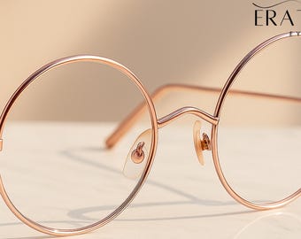 May include: Round eyeglasses with a thin rose gold metal frame. The glasses feature clear lenses, a high bridge, and adjustable nose pads. The brand name ERATIQUE EYEWEAR is printed in black text in the upper right corner of the image.