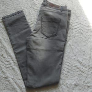 May include: Gray denim jeans with a classic five-pocket design. The jeans have a faded appearance and a straight leg cut. The back pocket features a subtle design. The jeans are laid flat on a textured white surface.
