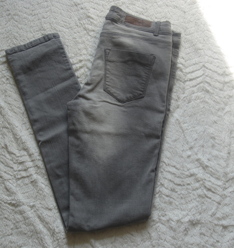 May include: Gray denim jeans with a classic five-pocket design. The jeans have a faded appearance and a straight leg cut. The back pocket features a subtle design. The jeans are laid flat on a textured white surface.