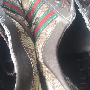 Puede incluir: Primer plano de zapatillas marrones con el logo de Gucci. Las zapatillas presentan un material de lona con el patr&oacute;n del logo de Gucci, detalles en cuero marr&oacute;n y un dise&ntilde;o de rayas verdes y rojas.