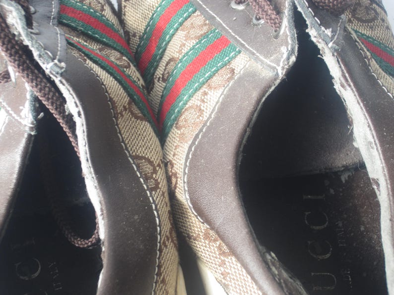 Puede incluir: Primer plano de zapatillas marrones con el logo de Gucci. Las zapatillas presentan un material de lona con el patr&oacute;n del logo de Gucci, detalles en cuero marr&oacute;n y un dise&ntilde;o de rayas verdes y rojas.
