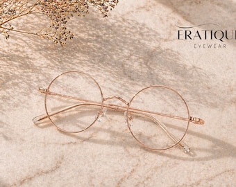 May include: Rose gold round eyeglasses with thin frames. The glasses are set against a light-colored, textured background. The brand name "ERATIQUE EYEWEAR" is displayed in the upper right corner.