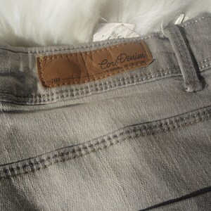 May include: Close-up of gray denim jeans with a brown leather label that reads "Core Denim" and "ZARA". The jeans have visible stitching and a belt loop. The fabric appears to be a light wash.