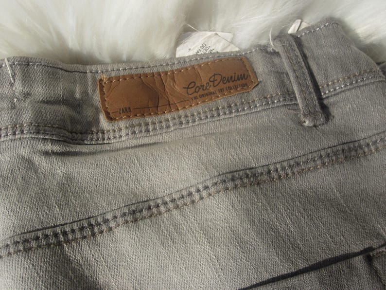 May include: Close-up of gray denim jeans with a brown leather label that reads "Core Denim" and "ZARA". The jeans have visible stitching and a belt loop. The fabric appears to be a light wash.
