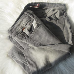 May include: Folded gray denim jeans with a classic five-pocket design. The jeans are a light gray color and appear to be made of a soft denim material. The jeans are folded neatly on a white, fluffy surface.