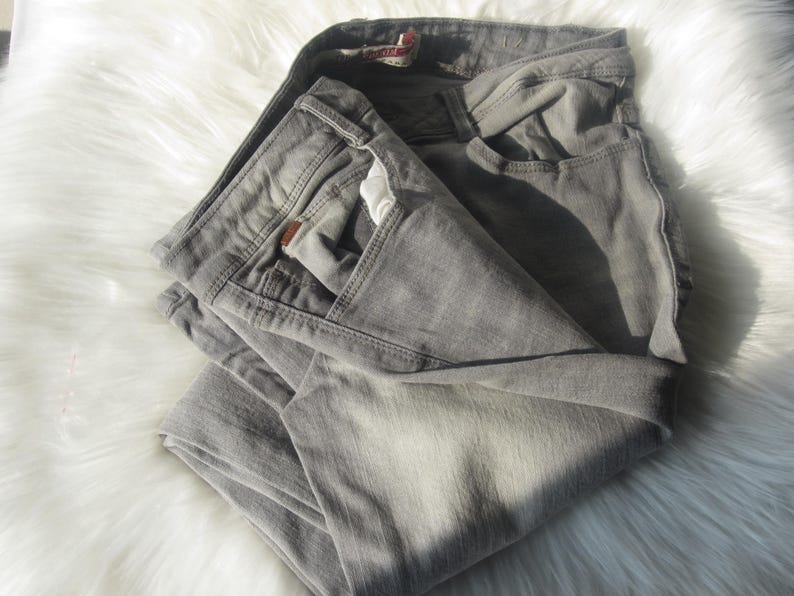 May include: Folded gray denim jeans with a classic five-pocket design. The jeans are a light gray color and appear to be made of a soft denim material. The jeans are folded neatly on a white, fluffy surface.