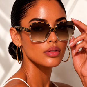 May include: Square-framed sunglasses featuring a brown and black leopard print pattern on the upper rim and temples, transitioning into a clear bottom frame. The lenses have a brown gradient tint. The text ERATIQUE EYEWEAR is visible.