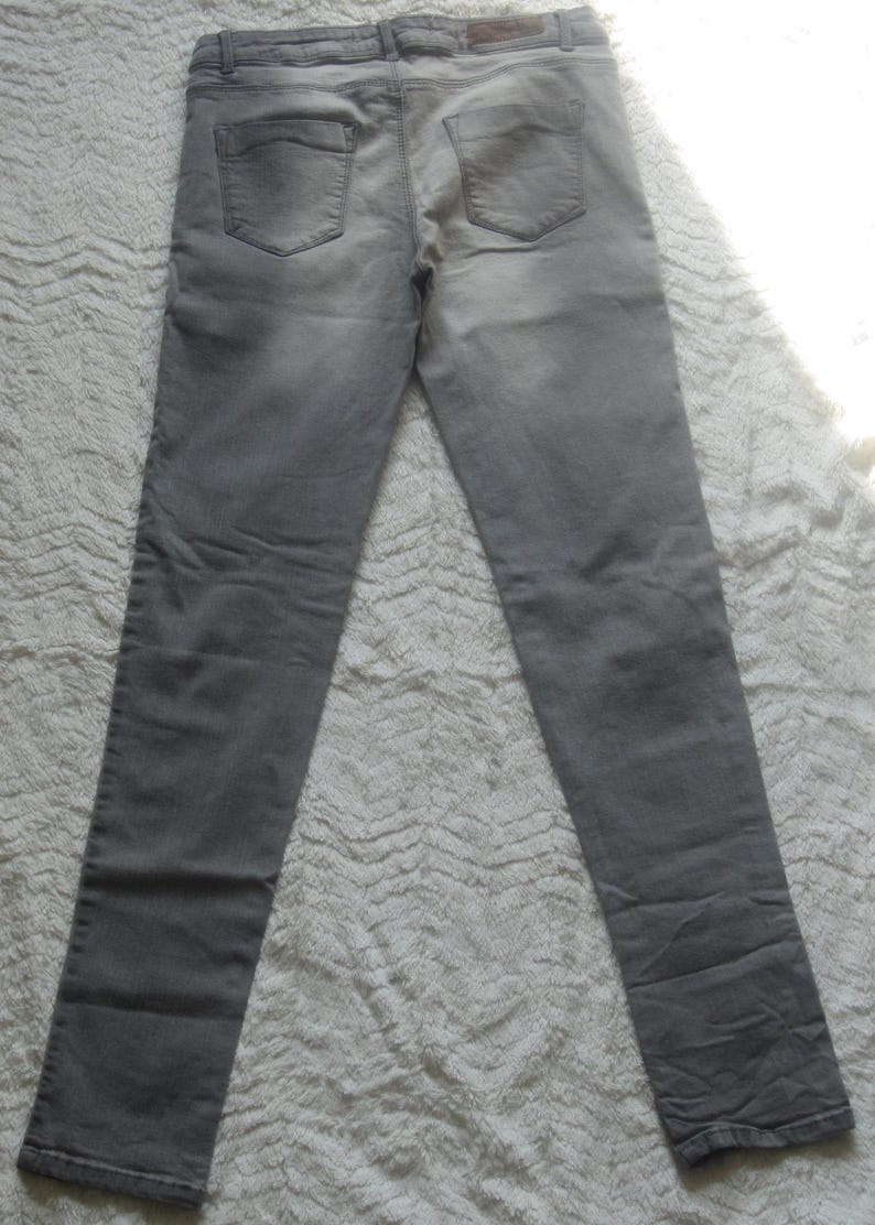 May include: A pair of gray denim jeans, laid flat, showcasing the back pockets and waistband. The jeans have a classic five-pocket design and a faded appearance. The fabric appears to be a medium-weight denim.