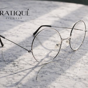 May include: Silver-framed round eyeglasses with clear lenses, resting on a marble surface. The glasses have thin temples and a bridge, with the brand name "ERATIQUE EYEWEAR" in the upper left corner.