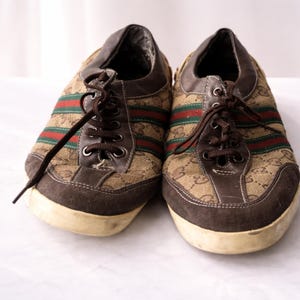 Puede incluir: Un par de zapatillas marrones con la parte superior de lona con un patr&oacute;n repetido. Las zapatillas tienen detalles de ante marr&oacute;n, rayas rojas y verdes y cordones marrones. Las suelas son blanquecinas.