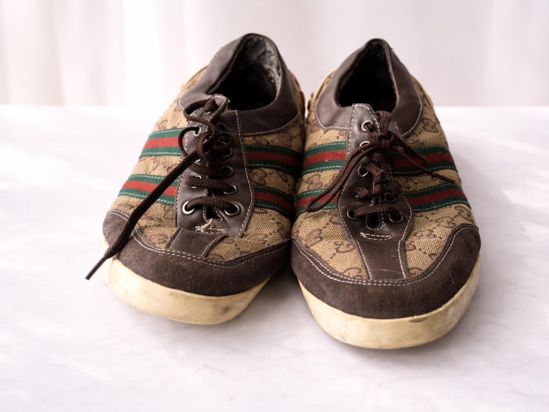 Puede incluir: Un par de zapatillas marrones con la parte superior de lona con un patr&oacute;n repetido. Las zapatillas tienen detalles de ante marr&oacute;n, rayas rojas y verdes y cordones marrones. Las suelas son blanquecinas.