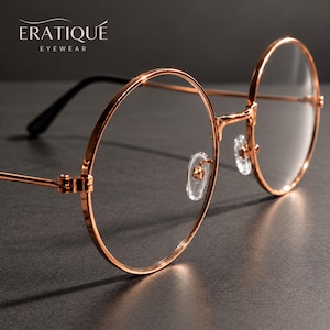 May include: Round eyeglasses with a polished rose gold metal frame and clear lenses. The temples feature black tips, and the bridge includes adjustable transparent nose pads. The text ERATIQUE EYEWEAR is visible in the upper left corner.