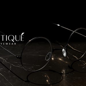 May include: Black round-frame eyeglasses with thin metal arms, resting on a dark marble surface. The eyeglasses are the focal point, with the brand name "ERATIQUE EYEWEAR" in white script above them. The background is black.