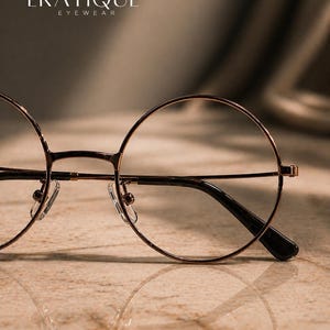May include: Close-up of round, wire-rimmed eyeglasses with a brown metal frame and black temples. The glasses are set against a neutral background, with the brand name "ERATIQUE EYEWEAR" visible at the top.